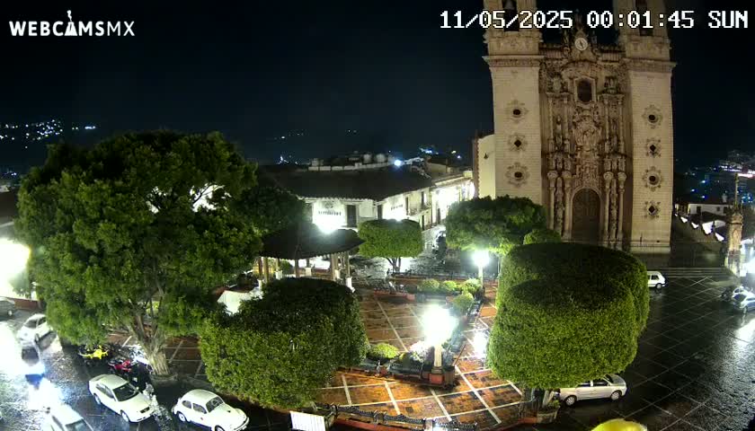 A nighttime view of a town square with a large church, numerous trees, and parked cars, under wet conditions.