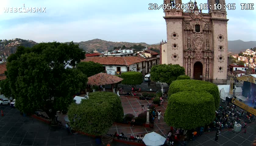 An outdoor plaza scene under a partly cloudy sky shows a large church, a gazebo, numerous trees, and many people gathered in the square.
