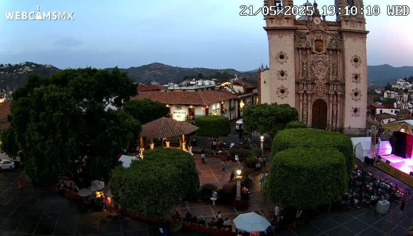 An outdoor plaza at dusk, featuring a large church, several buildings, lush green trees, and people gathered in the square under a twilight sky.