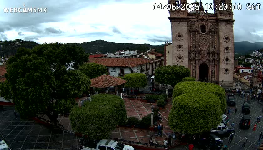 An overcast day reveals a town square with a large church, several vehicles, and lush green trees surrounding a paved plaza.