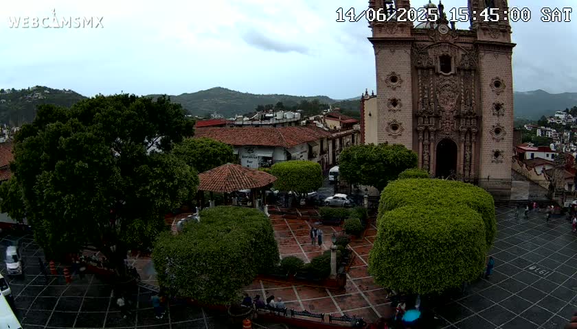 An overcast day reveals a town square with a large church, several vehicles, and lush green trees surrounding a paved plaza.