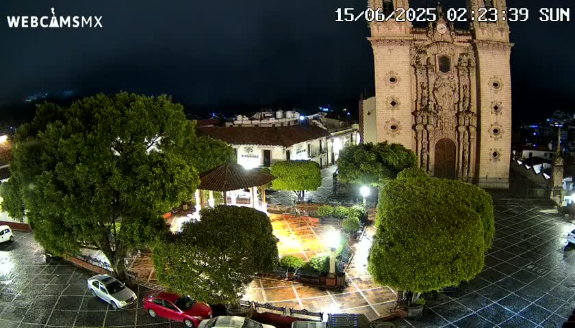 A nighttime view of a town square with a gazebo, lush green trees, a large church, and cars parked in the surrounding area, under a dark, possibly overcast sky.