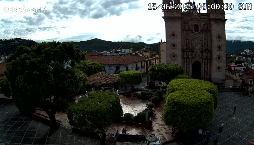 A town square with a large church, surrounded by lush green trees and shrubs under a cloudy sky.