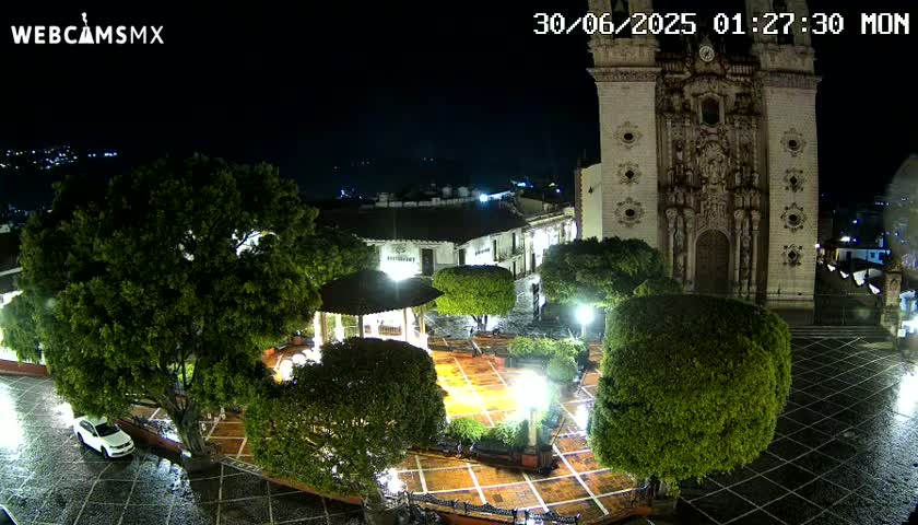 A nighttime view of a town square, illuminated by lights, featuring a large church and lush green trees, with a light rain falling on wet cobblestones.