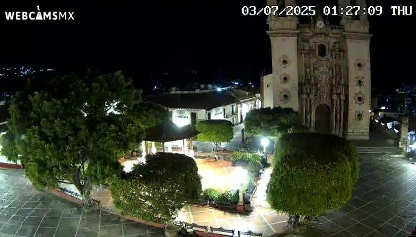 A nighttime view of a town square with a large church,  trees, and a gazebo, under a clear, dark sky.