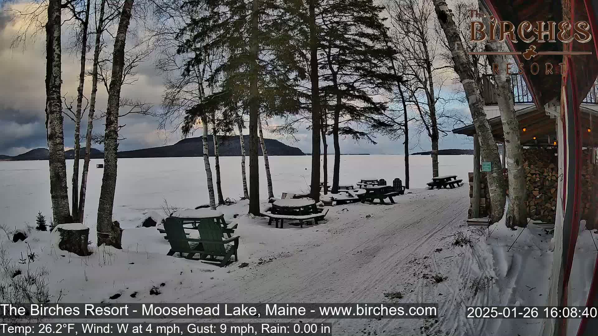 A snow-covered lakeside patio with picnic tables and chairs is situated near a building under a cloudy sky.