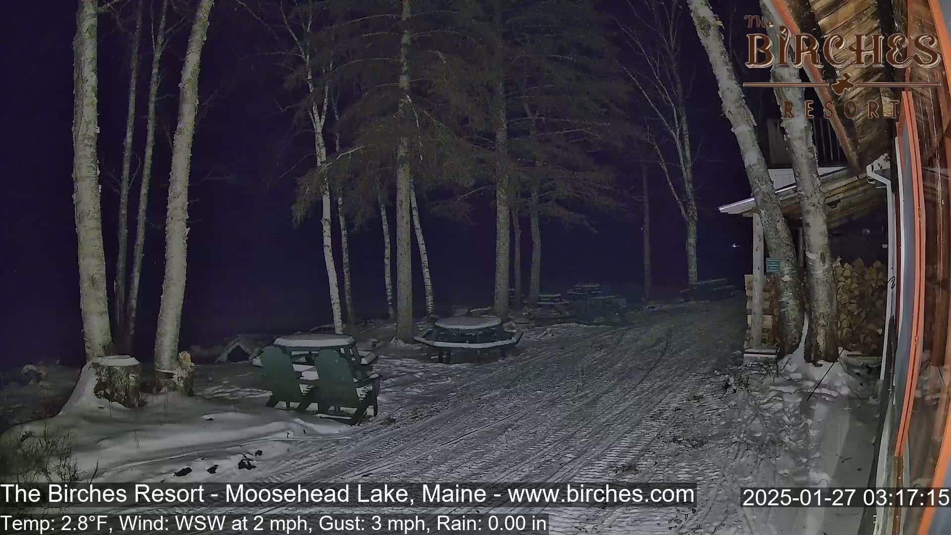 A snow-covered outdoor area at night, with several empty picnic tables under birch trees near a building, shows a light wind and a temperature of 2.8°F.