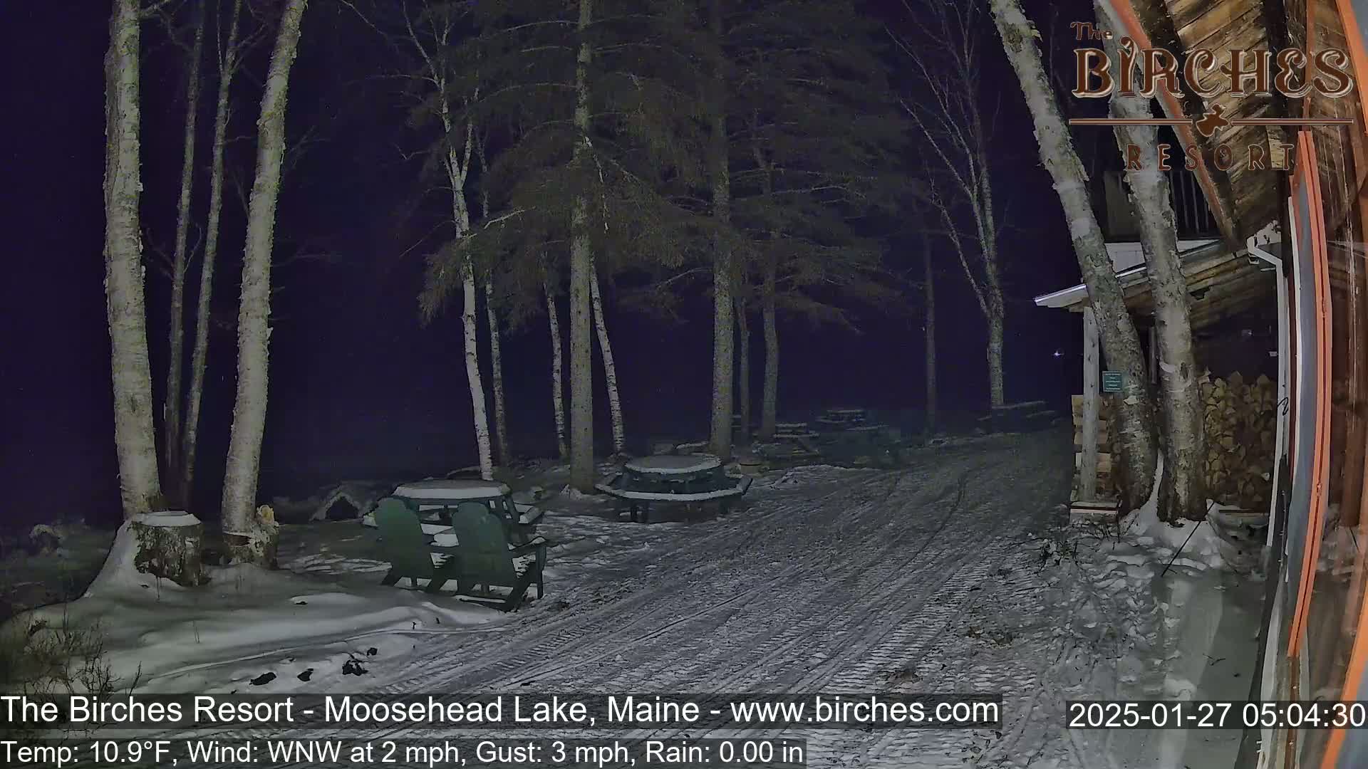 A snow-covered outdoor area at night, illuminated by faint light, shows several picnic tables and chairs near a building under a dark sky with a light wind.