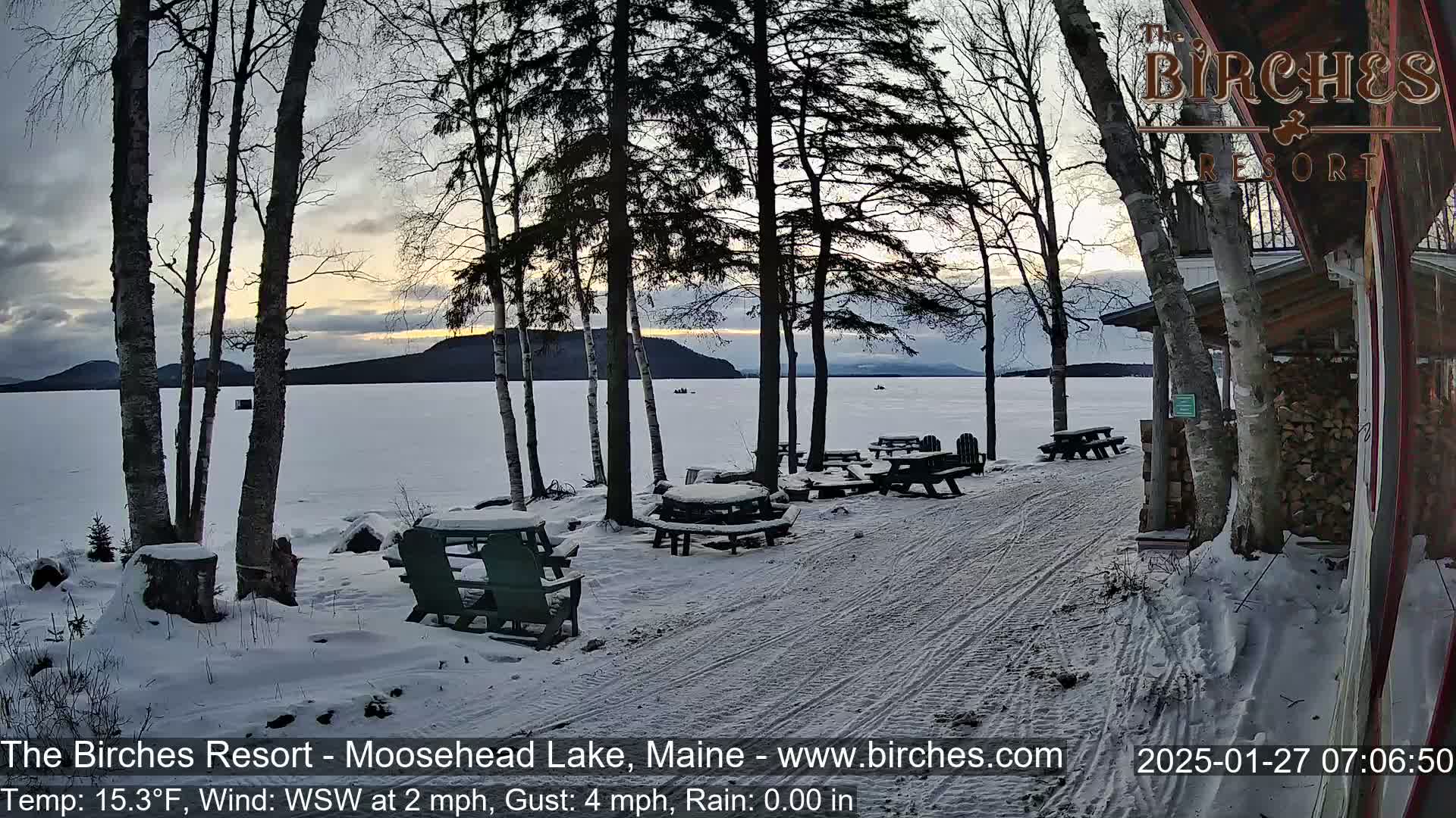 A snow-covered lakeside patio with picnic tables and chairs overlooks a frozen lake at sunset under a cloudy sky.