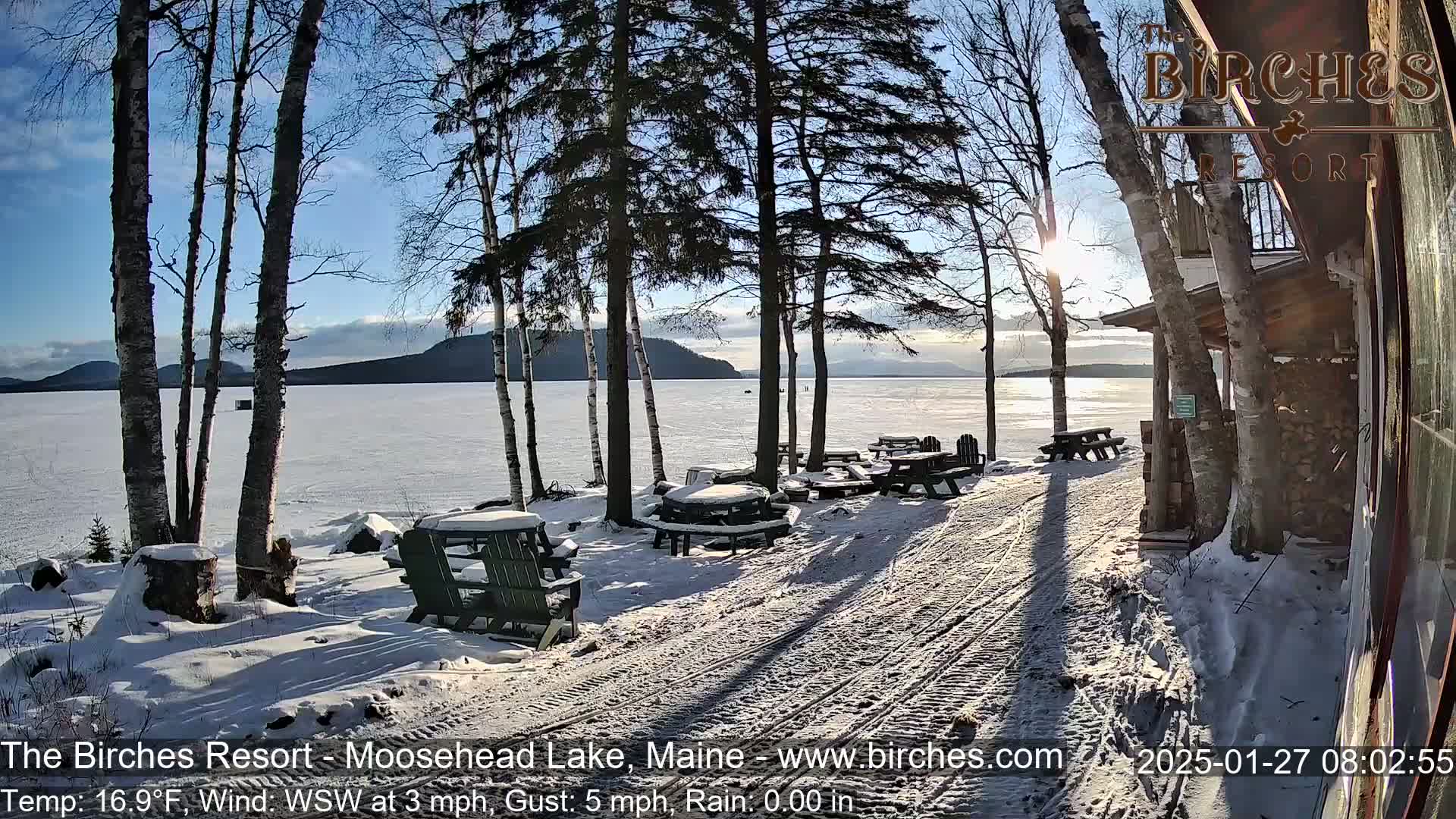 A snow-covered lakeside scene with picnic tables and a building, under a sunny, partly cloudy winter sky.