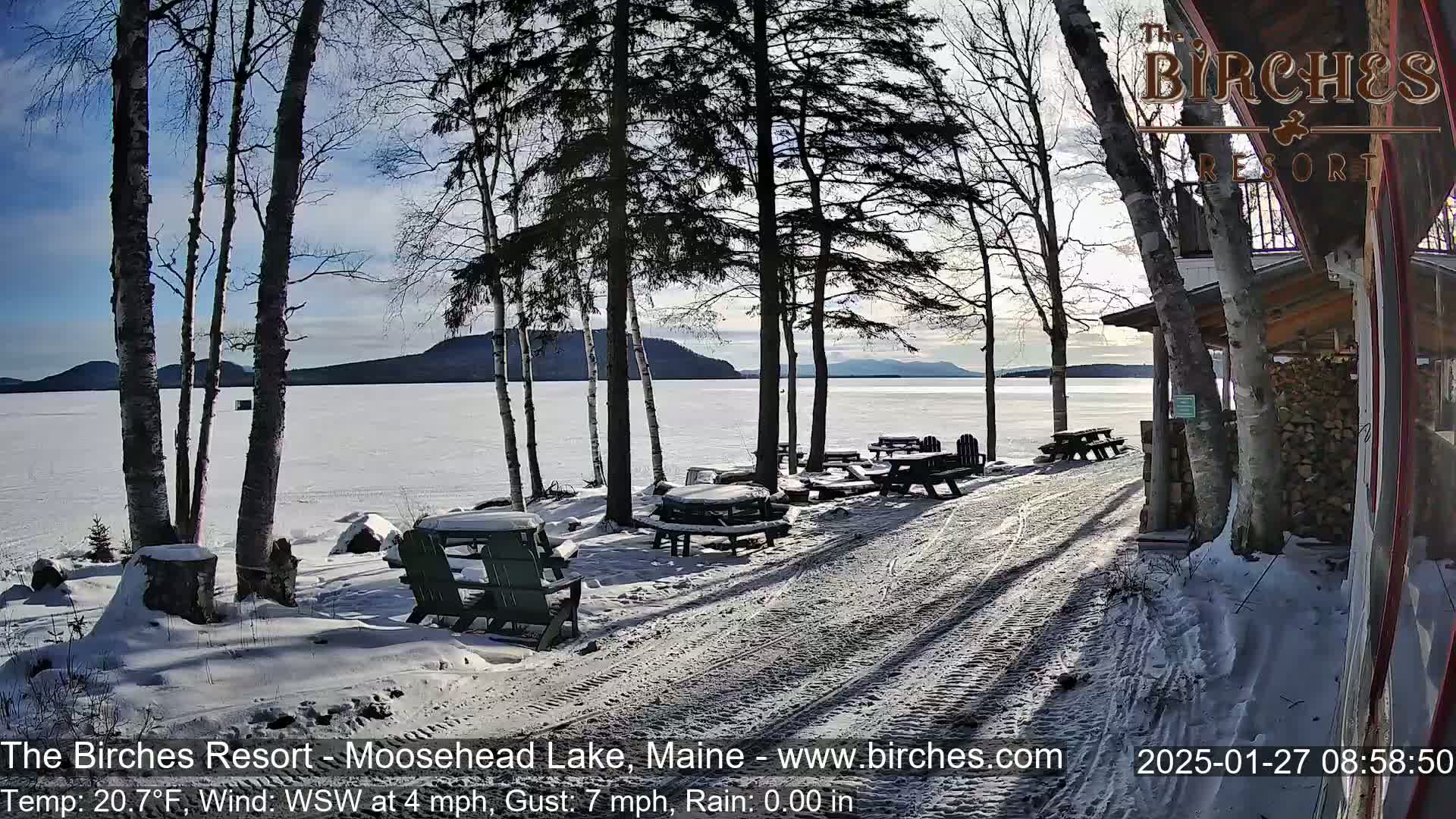 A snow-covered lakeside patio with picnic tables and chairs, overlooking a frozen lake under a mostly sunny sky.