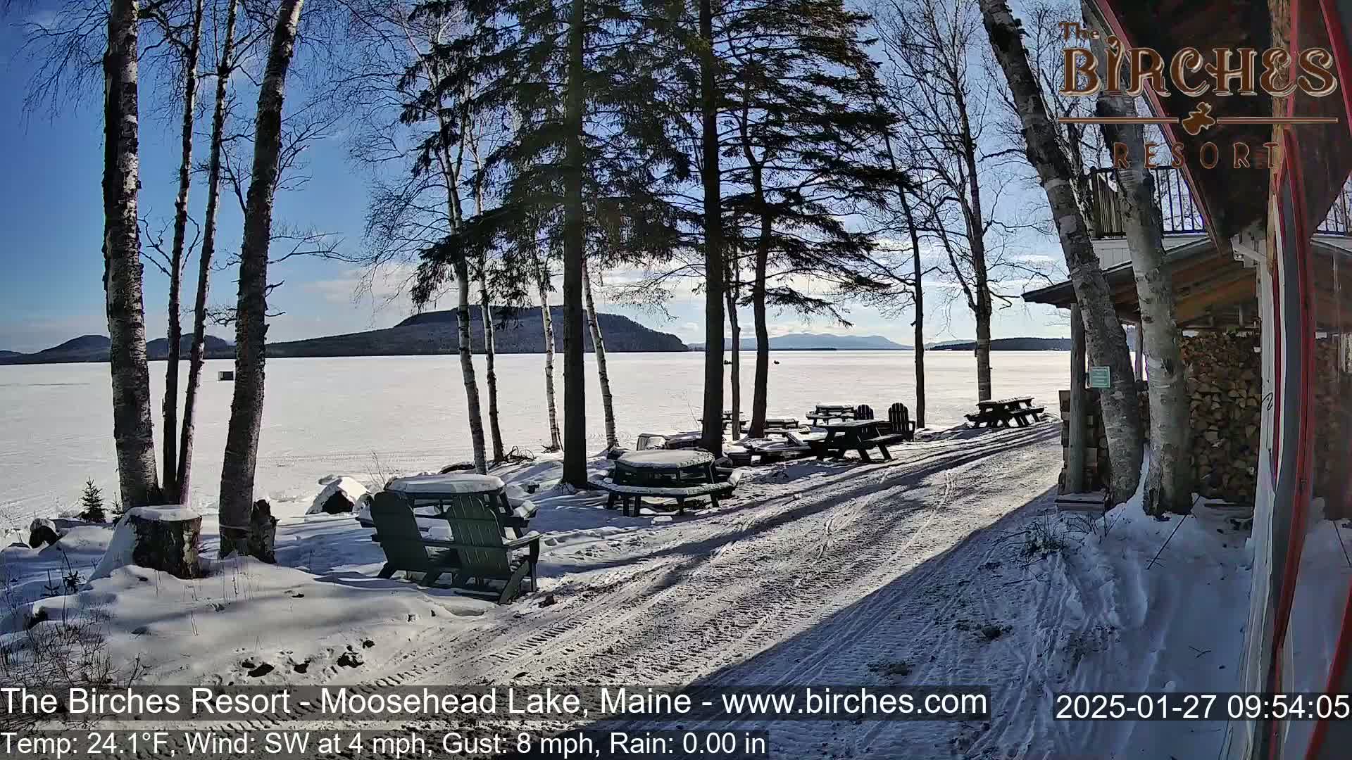 A snow-covered lakeside area with picnic tables and chairs under a sunny winter sky.