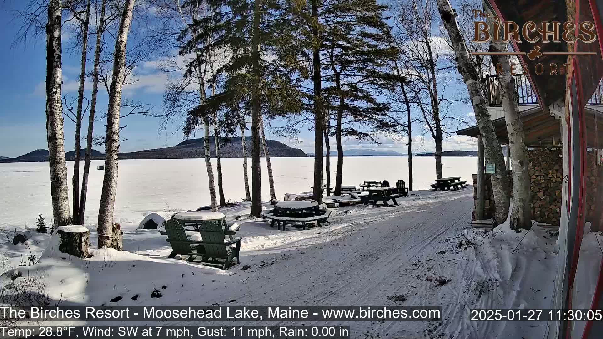 A snow-covered lakeside patio with picnic tables and Adirondack chairs overlooks a frozen lake under a sunny sky.