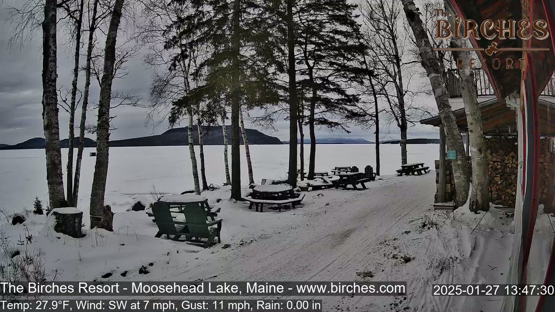 A snow-covered lakeside area with picnic tables and trees under a gray sky, with a southwest wind at 7 mph and gusts up to 11 mph.