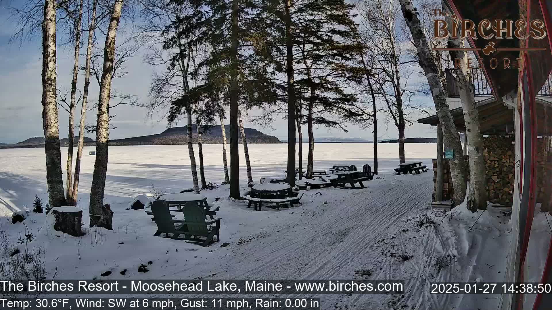 A snow-covered lakeside scene with picnic tables and Adirondack chairs near a building, under a partly sunny sky.