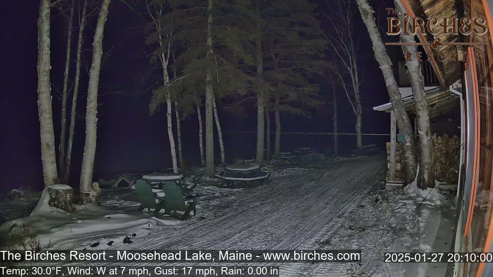A snow-covered path at night leads to picnic tables under birch trees near a building, with a 30°F temperature and a westerly wind of 7 mph gusting to 17 mph.