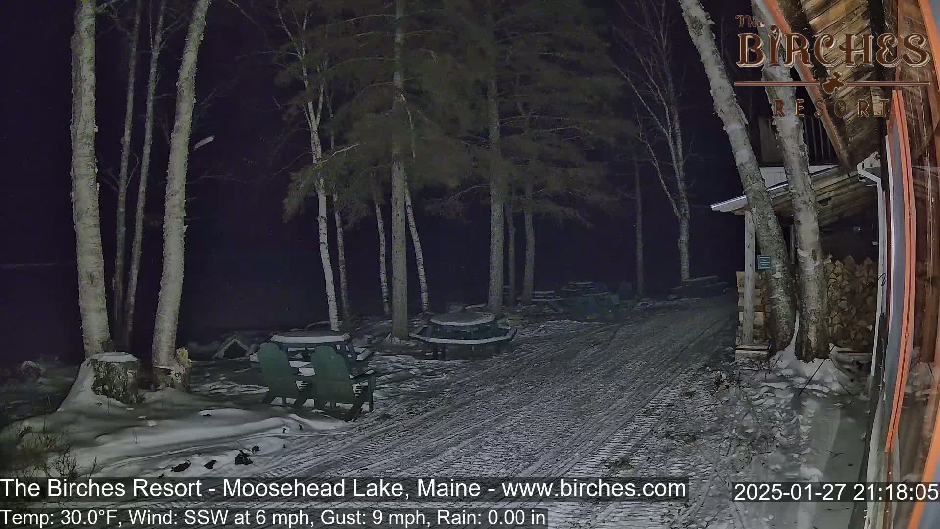 A snow-covered path at night leads to picnic tables and a building under a dark sky with a south-southwesterly wind.