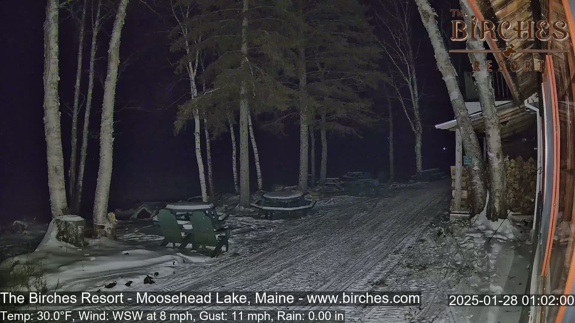 A snow-covered pathway at night leads to a building next to several picnic tables and trees under a dark sky with a WSW wind at 8 mph and gusts up to 11 mph.
