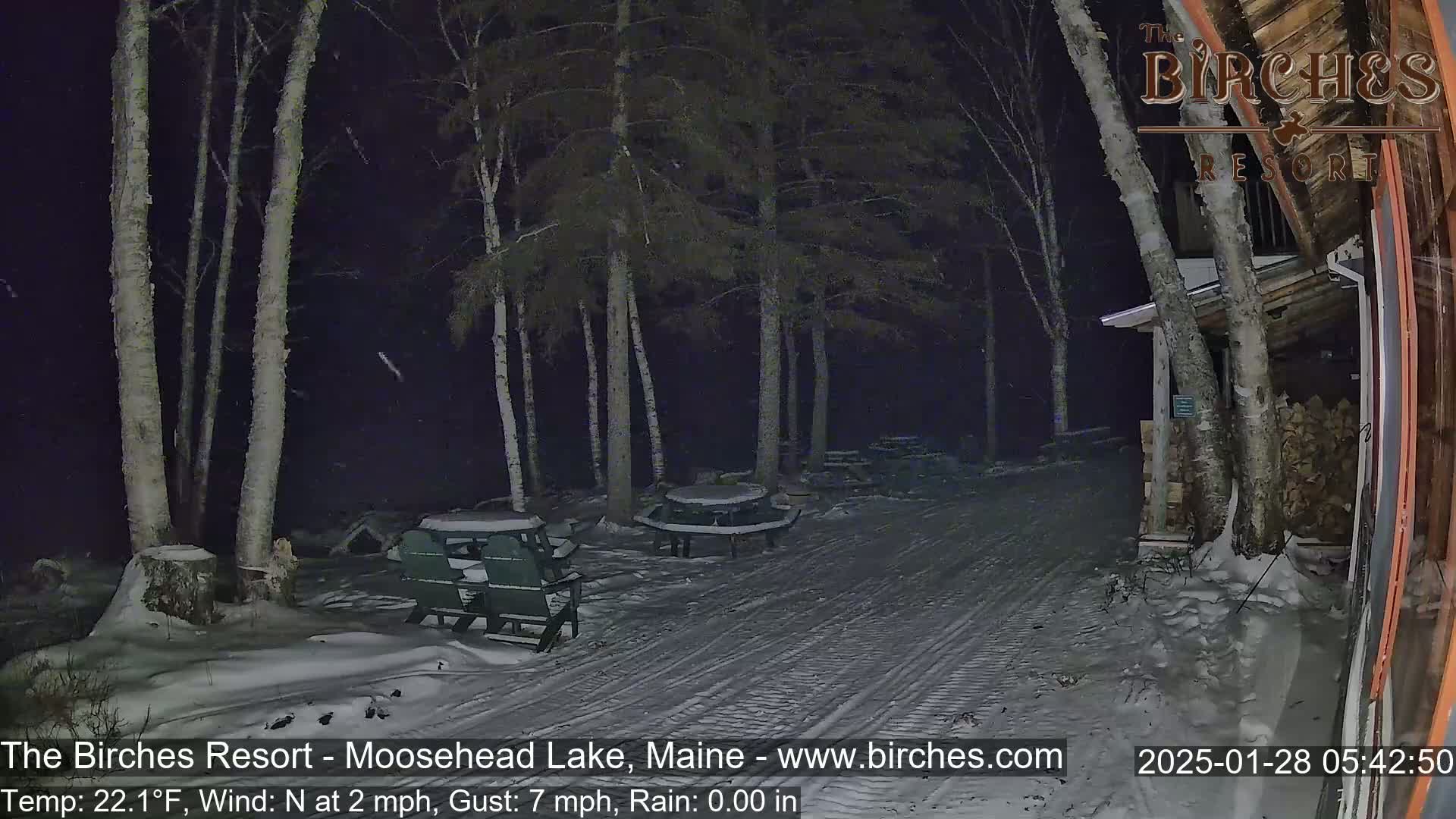 A snow-covered path at night, illuminated by ambient light, leads past picnic tables and chairs to a wooden building under a dark, lightly snowing sky.