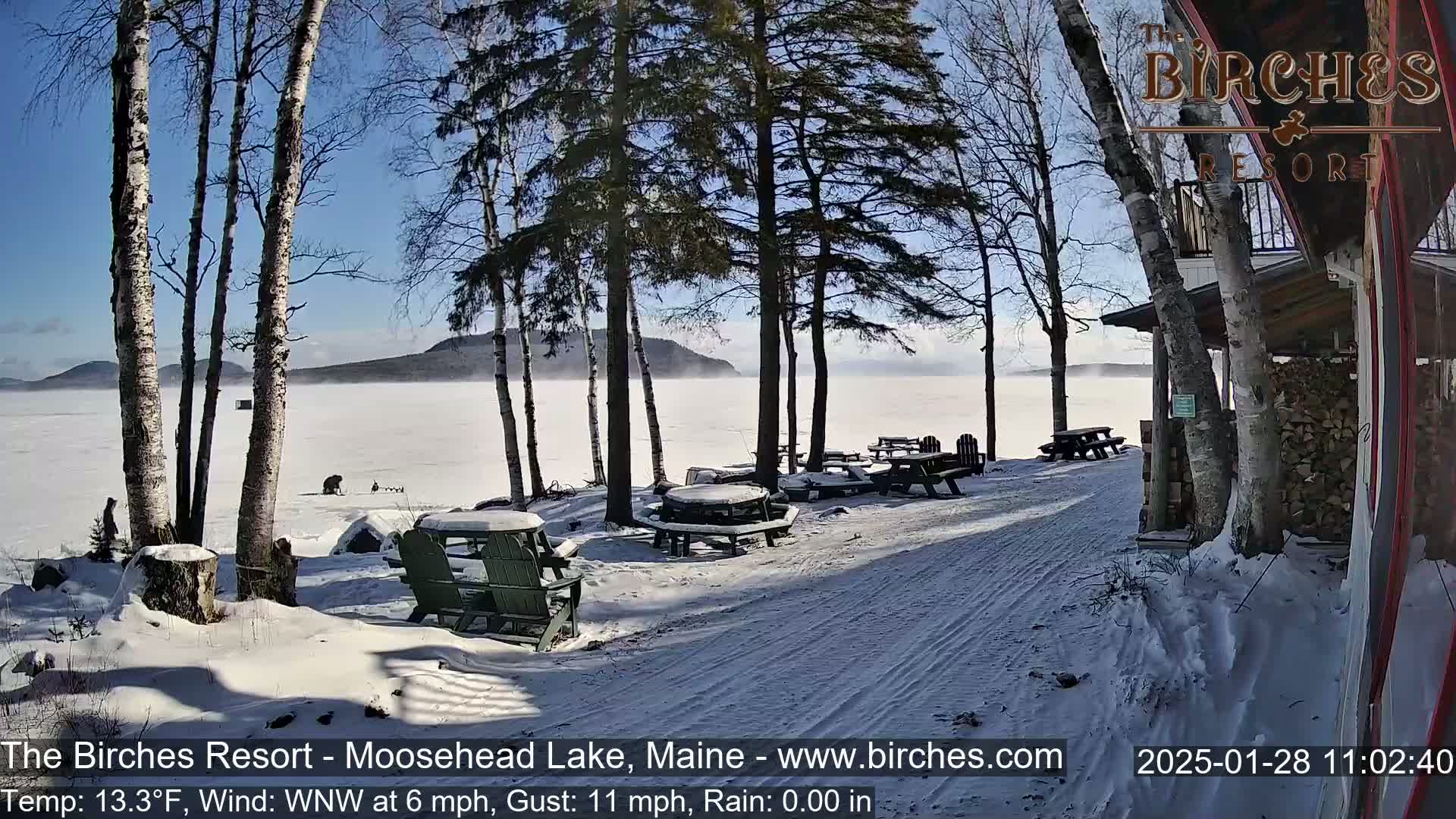 A snow-covered lakeside area with picnic tables and chairs, several birch trees, and a partially visible building, all under a sunny, cold winter sky with a light fog over the lake.