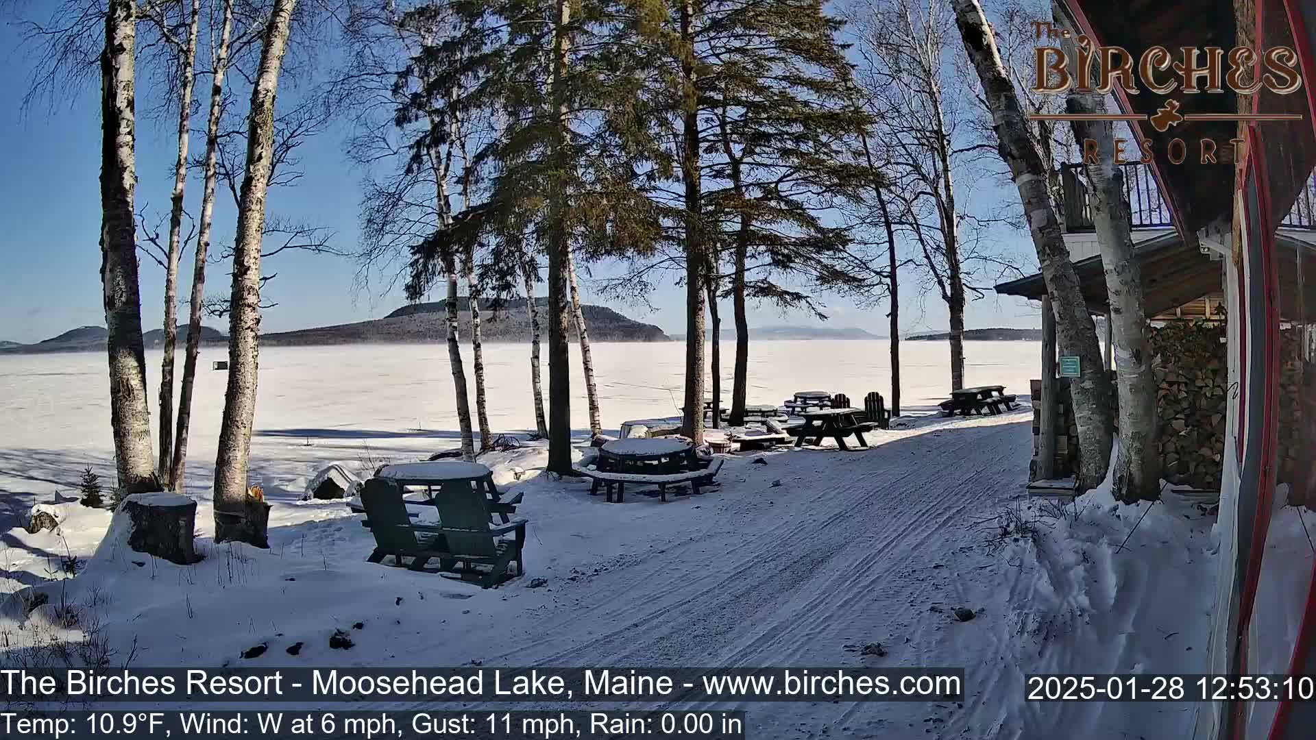 A snow-covered lakeside patio with picnic tables and chairs is visible, overlooking a frozen lake under a clear, sunny sky.