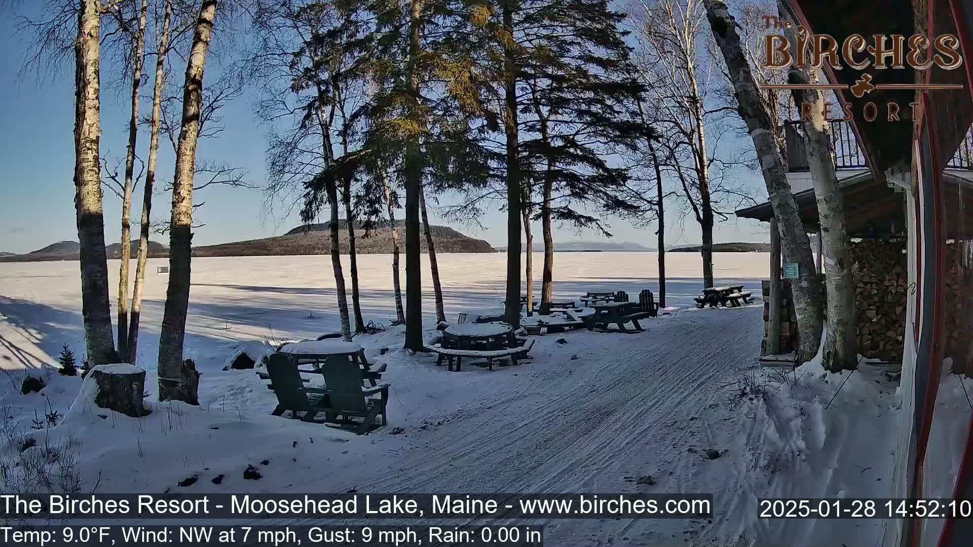 A snow-covered lakeside path leads to picnic tables under birch and pine trees, with a frozen lake and distant mountains visible under a sunny sky.