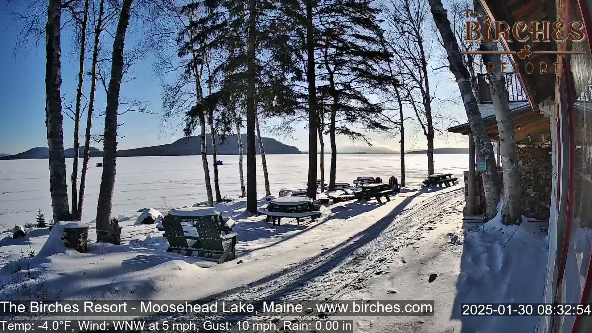 A snow-covered lakeside patio with picnic tables and chairs overlooks a frozen lake under a sunny, cold sky.