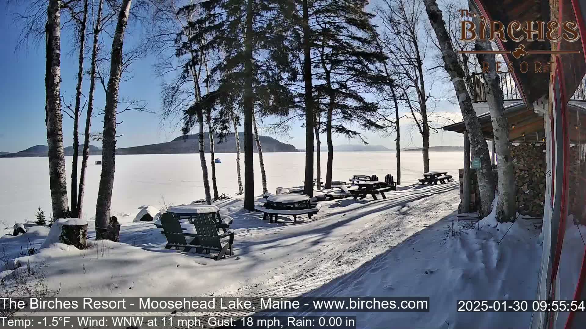 A snow-covered lakeside patio with picnic tables and chairs is seen under a sunny winter sky, with a frozen lake and distant mountains in the background.