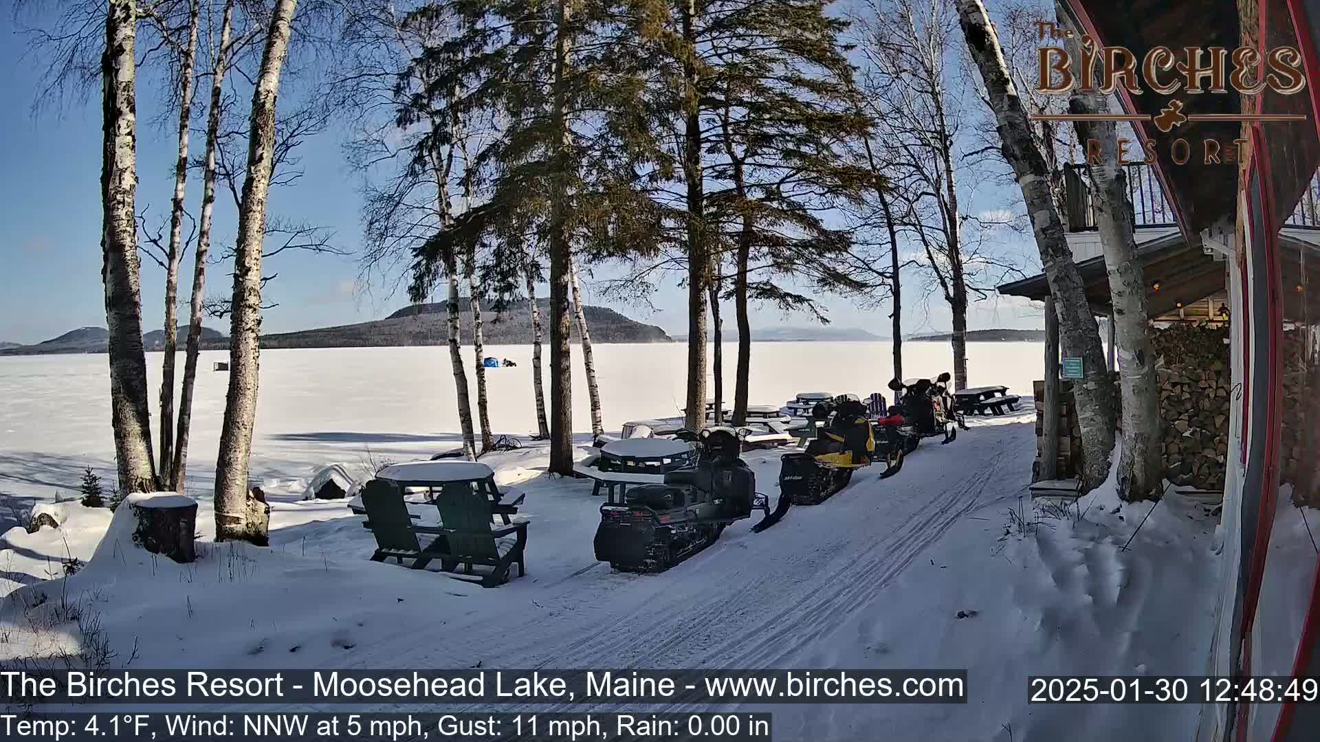 A sunny, snowy lakeside scene shows snowmobiles parked near a building and picnic tables, with a frozen lake and distant mountains in the background.