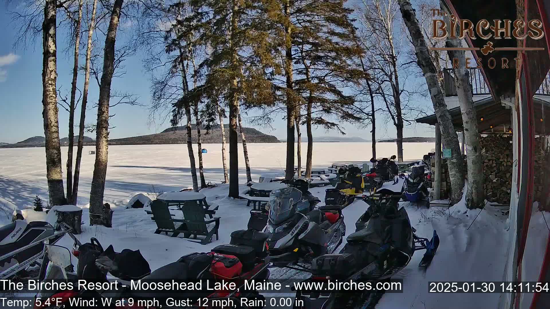Several snowmobiles are parked in the snow near a lakeside building on a sunny, cold day with a frozen lake in the background.