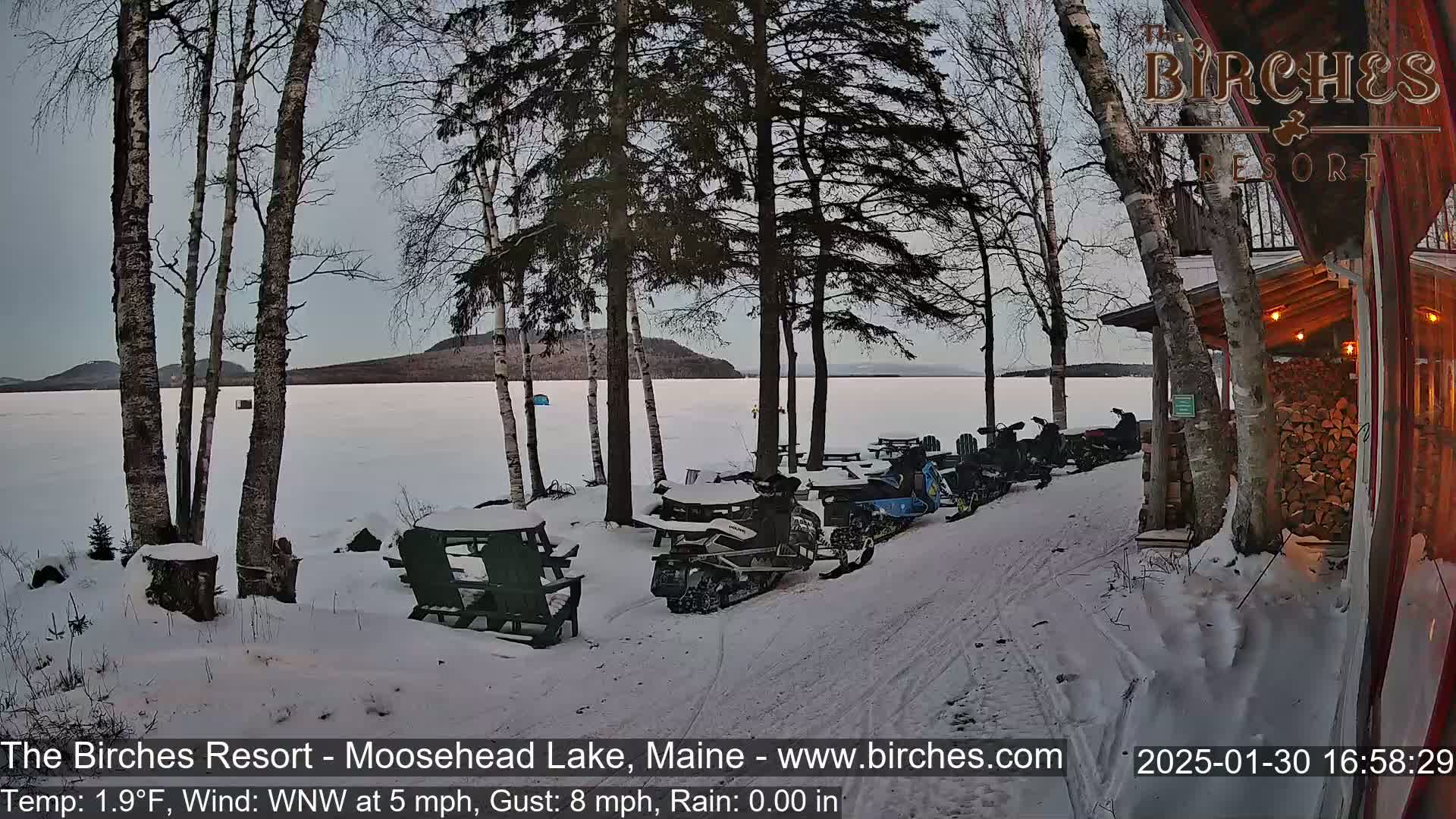 Several snowmobiles are parked near a lakeside cabin in a snowy landscape under a cloudy sky.