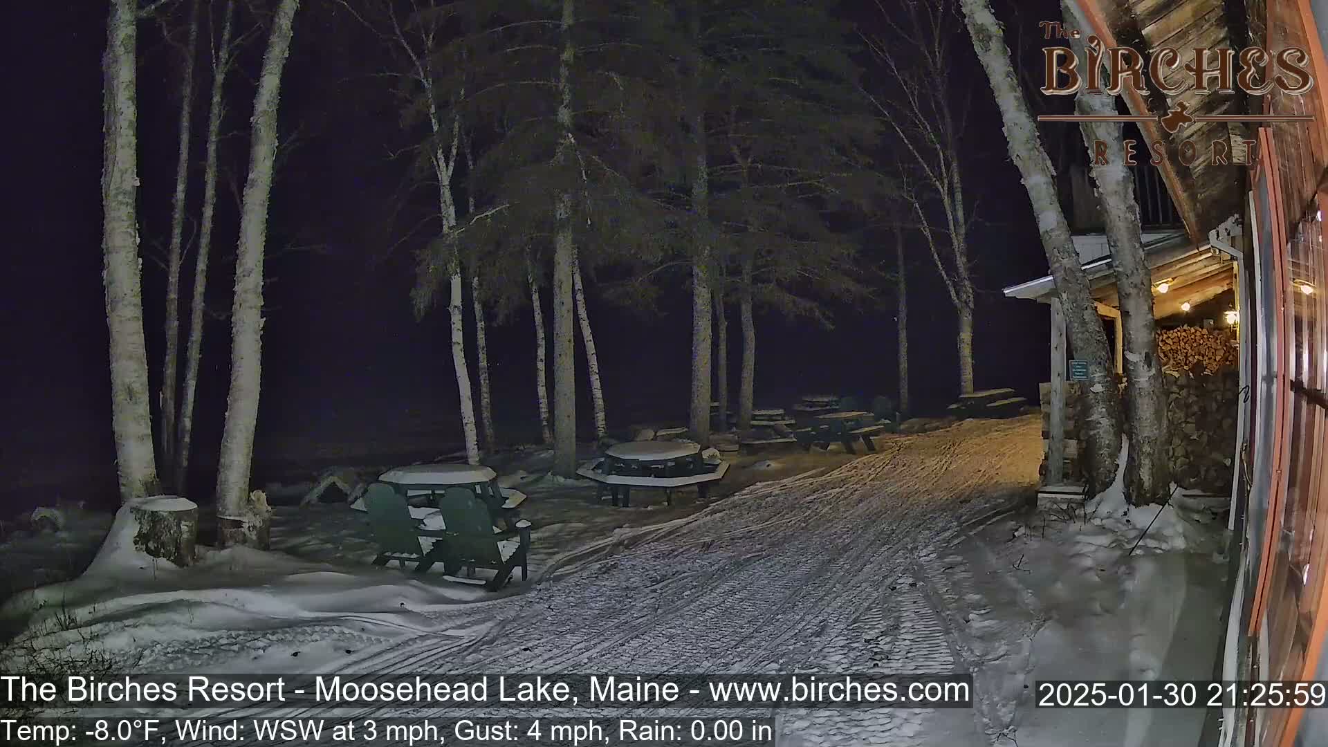 A snow-covered outdoor area at night, illuminated by lights, shows several picnic tables and chairs under tall birch trees near a building with a woodpile.