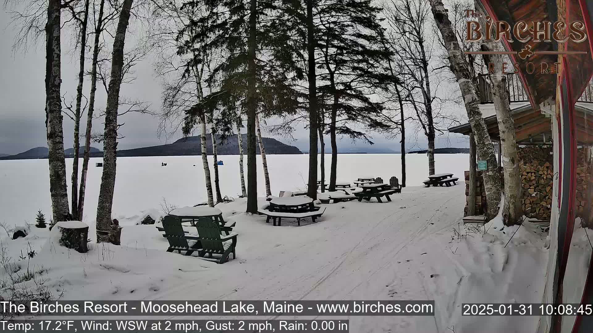 Snow-covered picnic tables sit near a snow-covered lake shore under a cloudy sky, with bare trees and a wooden building in the background.
