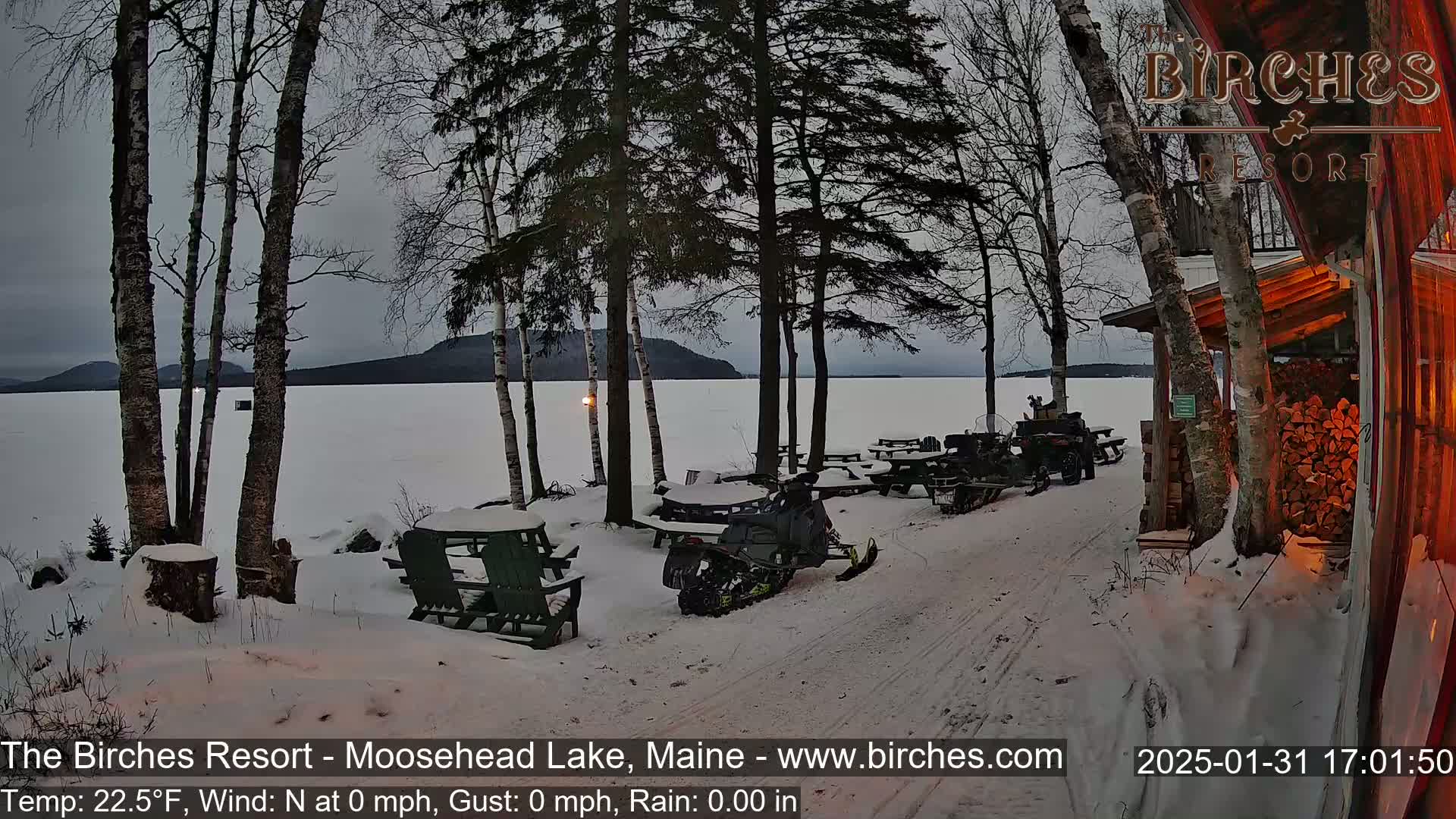 A snow-covered lakeside scene at dusk shows snowmobiles parked near picnic tables and a woodpile by a building, with calm weather and a frozen lake in the background.