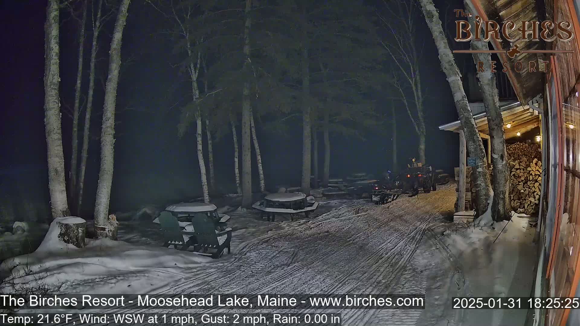 A snow-covered outdoor area at night, lit by artificial light, shows picnic tables, chairs, and a snowmobile parked near a building with a woodpile under a dark, partly cloudy sky.