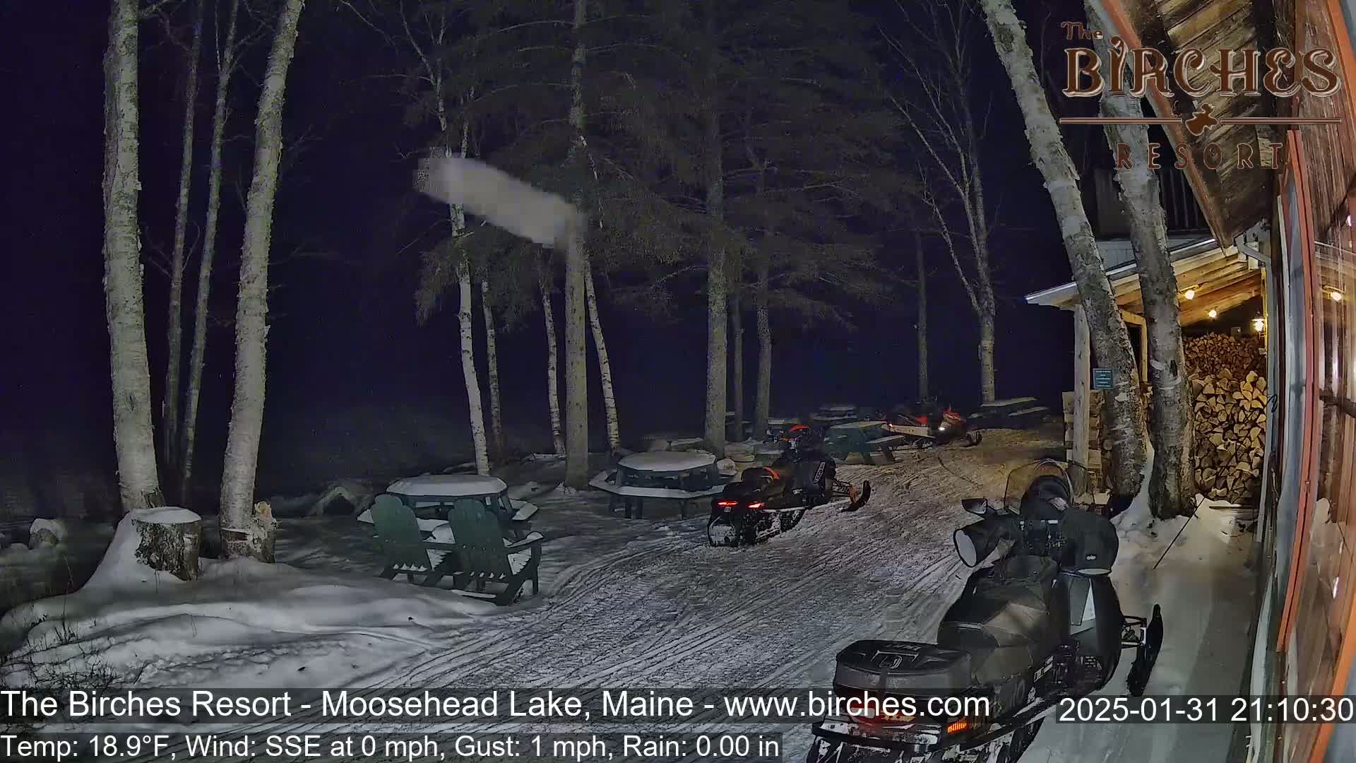 Snow-covered picnic tables and snowmobiles are visible at night in a snow-covered outdoor area near birch trees, under a dark sky with light snow falling.