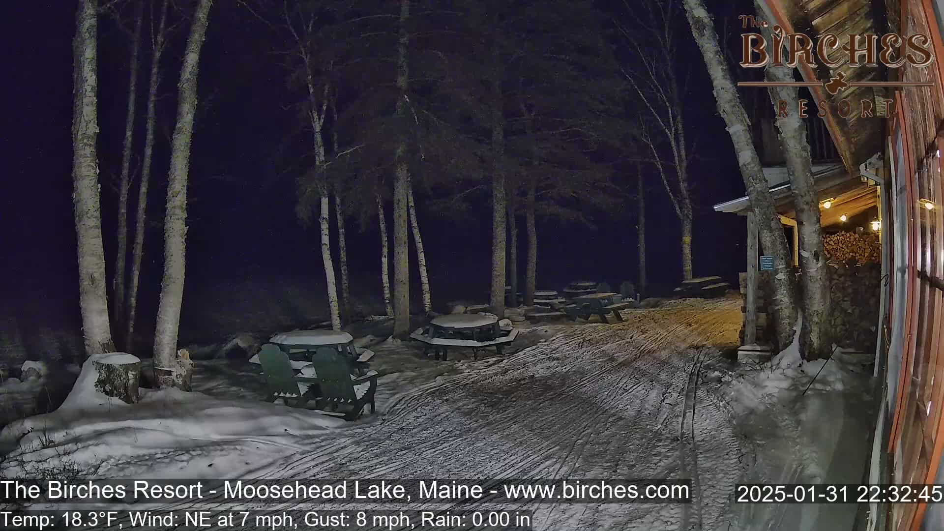 A snow-covered outdoor patio area with picnic tables and chairs is seen at night, under a dark sky with a northeast wind.