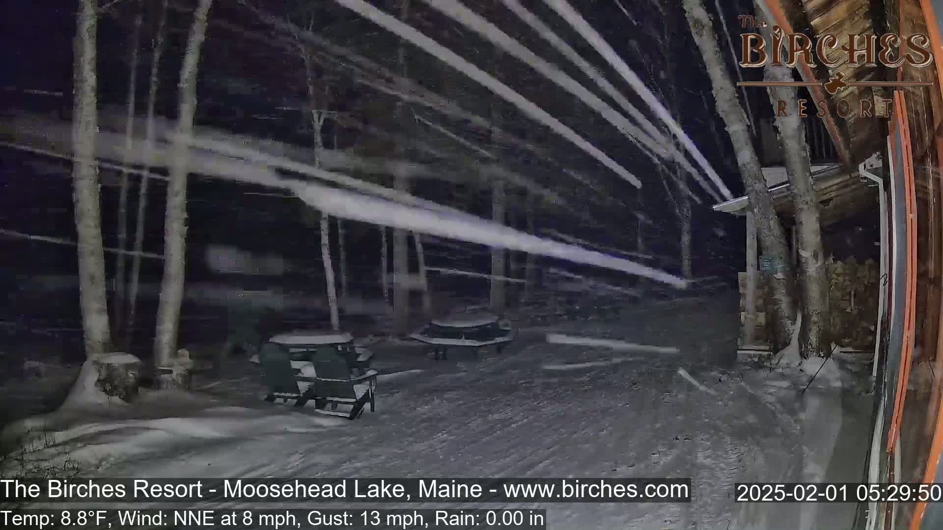Snow blows horizontally through a snow-covered outdoor area at night, past several chairs and a table, near a building and trees.