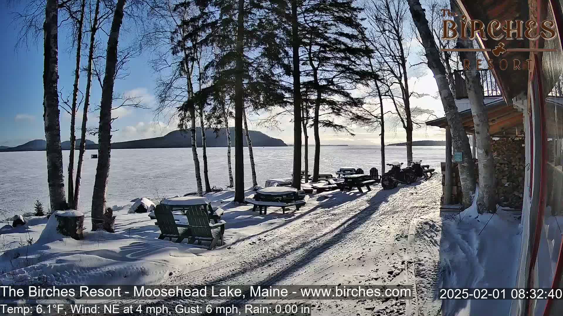 A snow-covered lakeside picnic area with several empty tables and chairs, situated next to a building and some snowmobiles, under a mostly sunny sky.