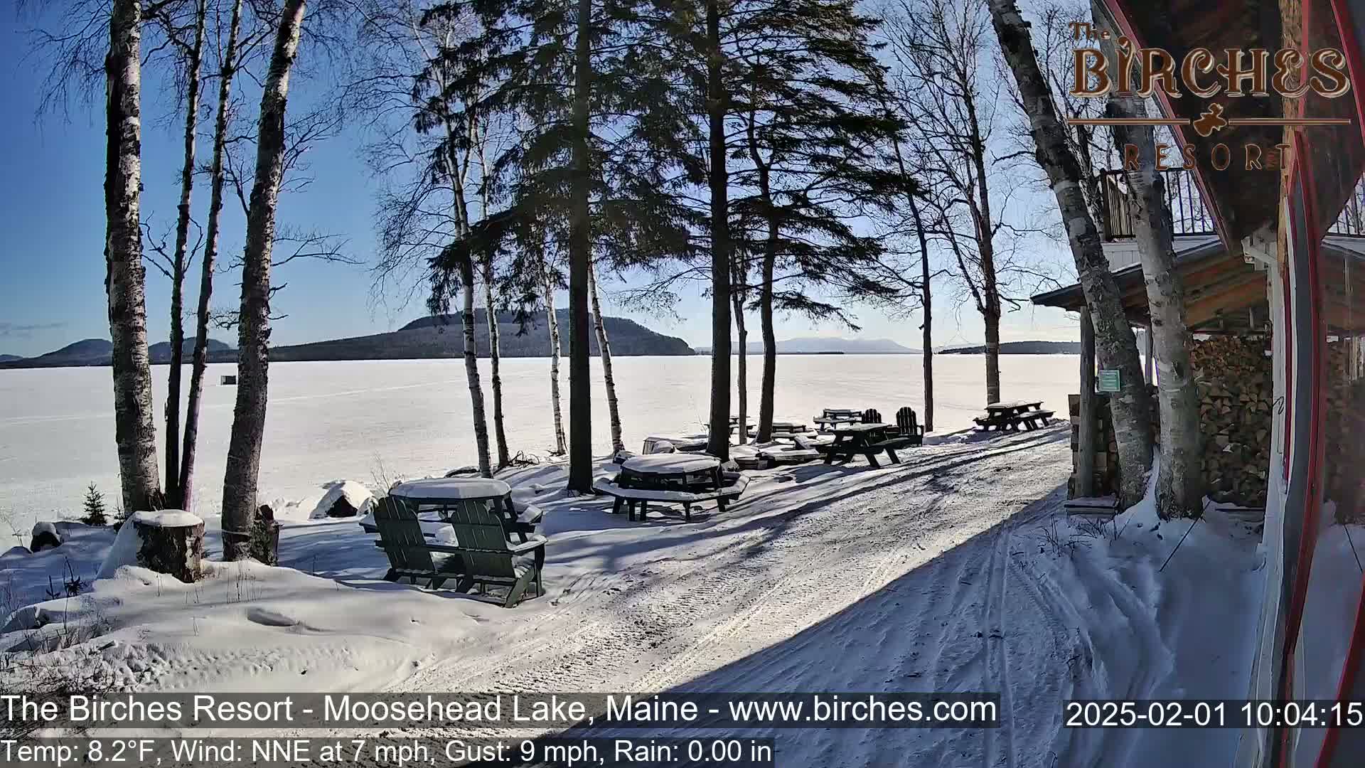 A snow-covered lakeside scene with picnic tables and Adirondack chairs under a sunny sky, featuring a frozen lake and distant mountains.