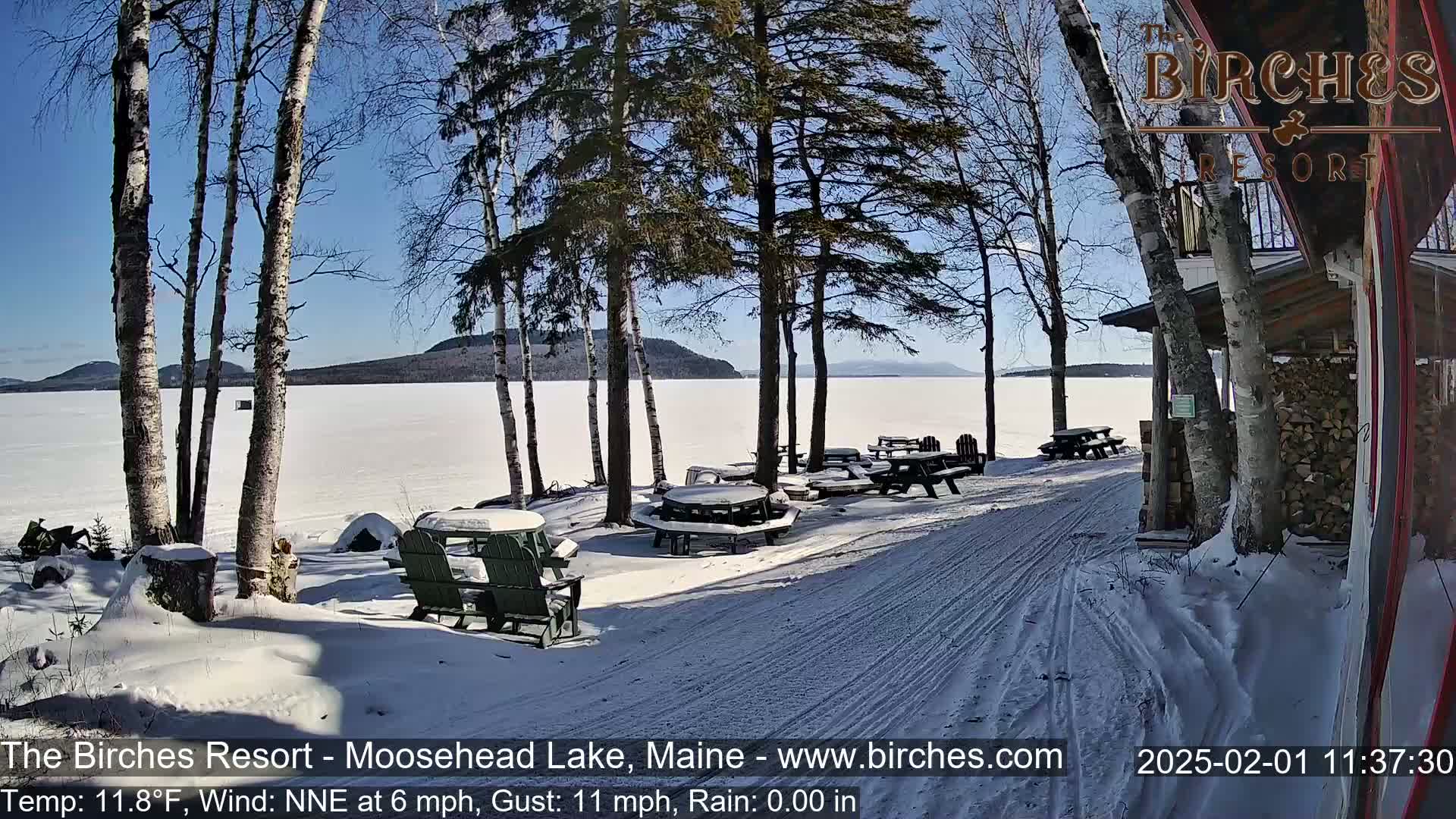 A snow-covered lakeside patio with picnic tables and chairs, under a sunny sky, overlooks a frozen lake and distant hills.