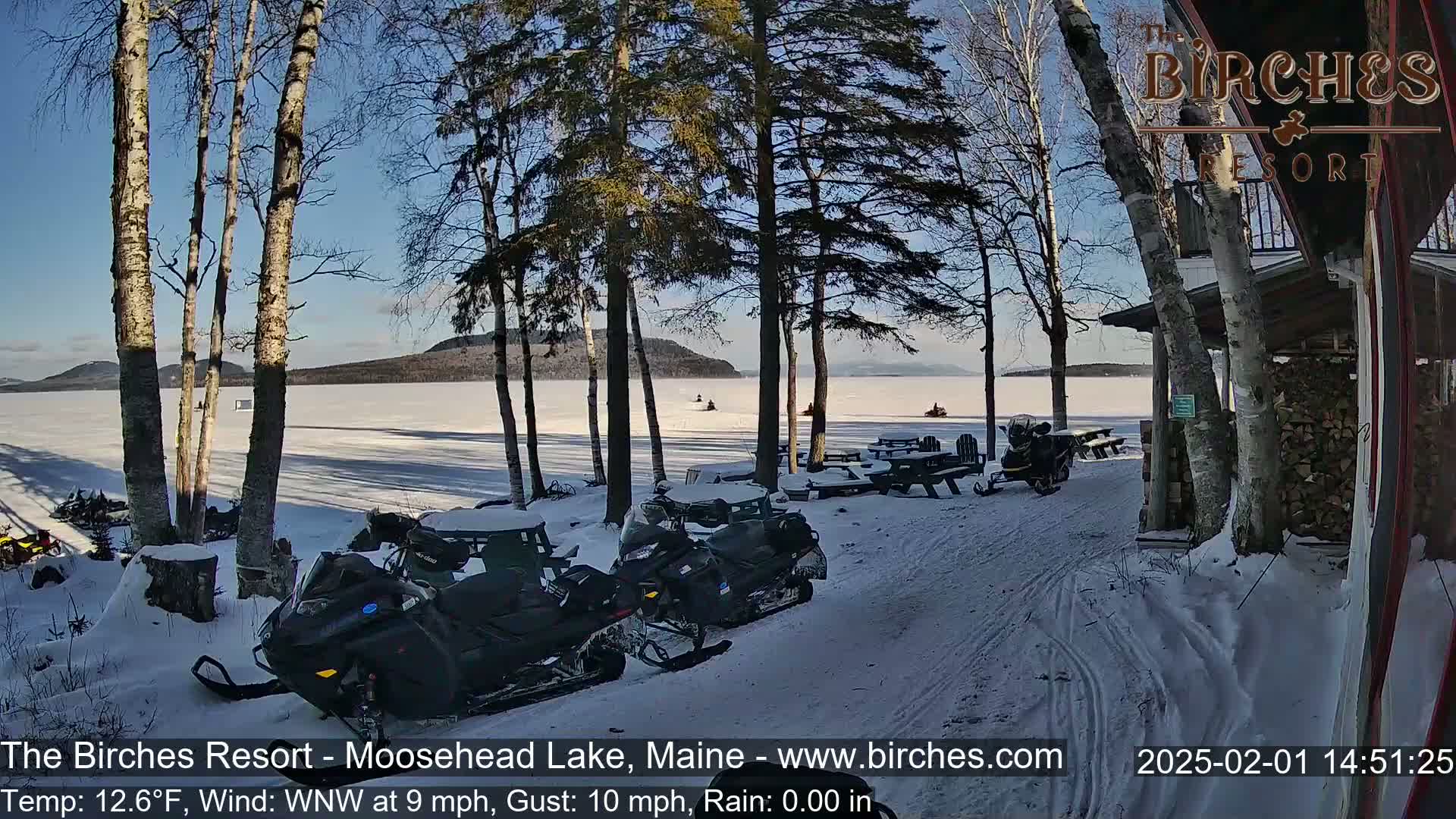 Several snowmobiles are parked near a snow-covered lakeshore under a sunny, cold sky.