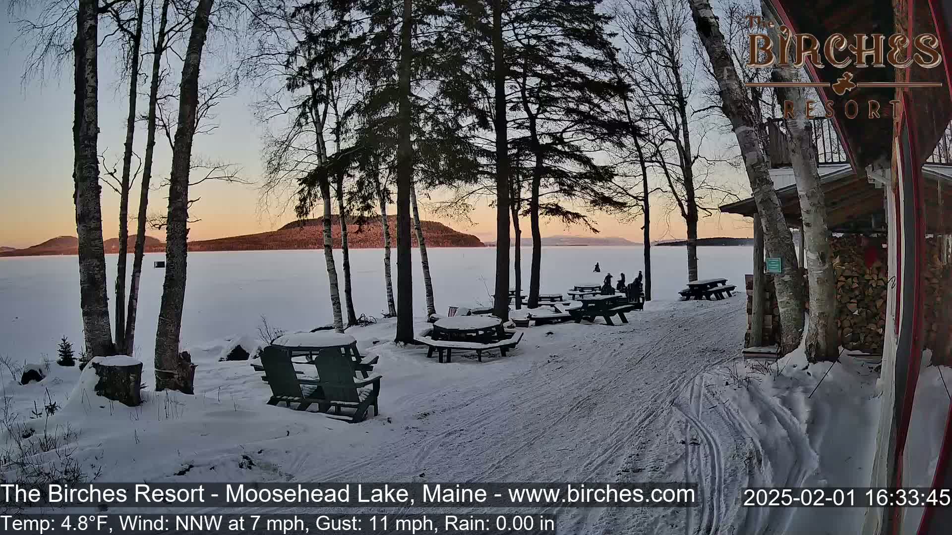 A snow-covered lakeside picnic area with several empty tables and chairs sits under a twilight sky next to a building, with a frozen lake in the background.