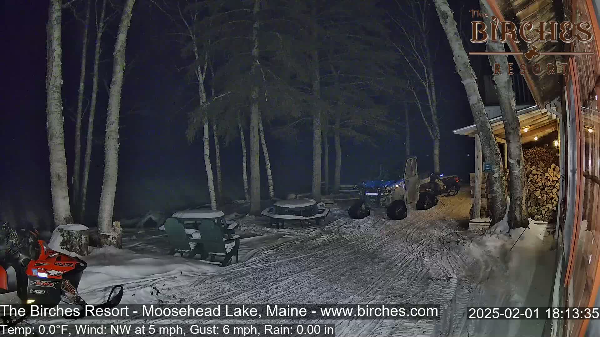 A snow-covered outdoor area at night, illuminated by artificial light, shows several snowmobiles parked near picnic tables and a building with a woodpile.