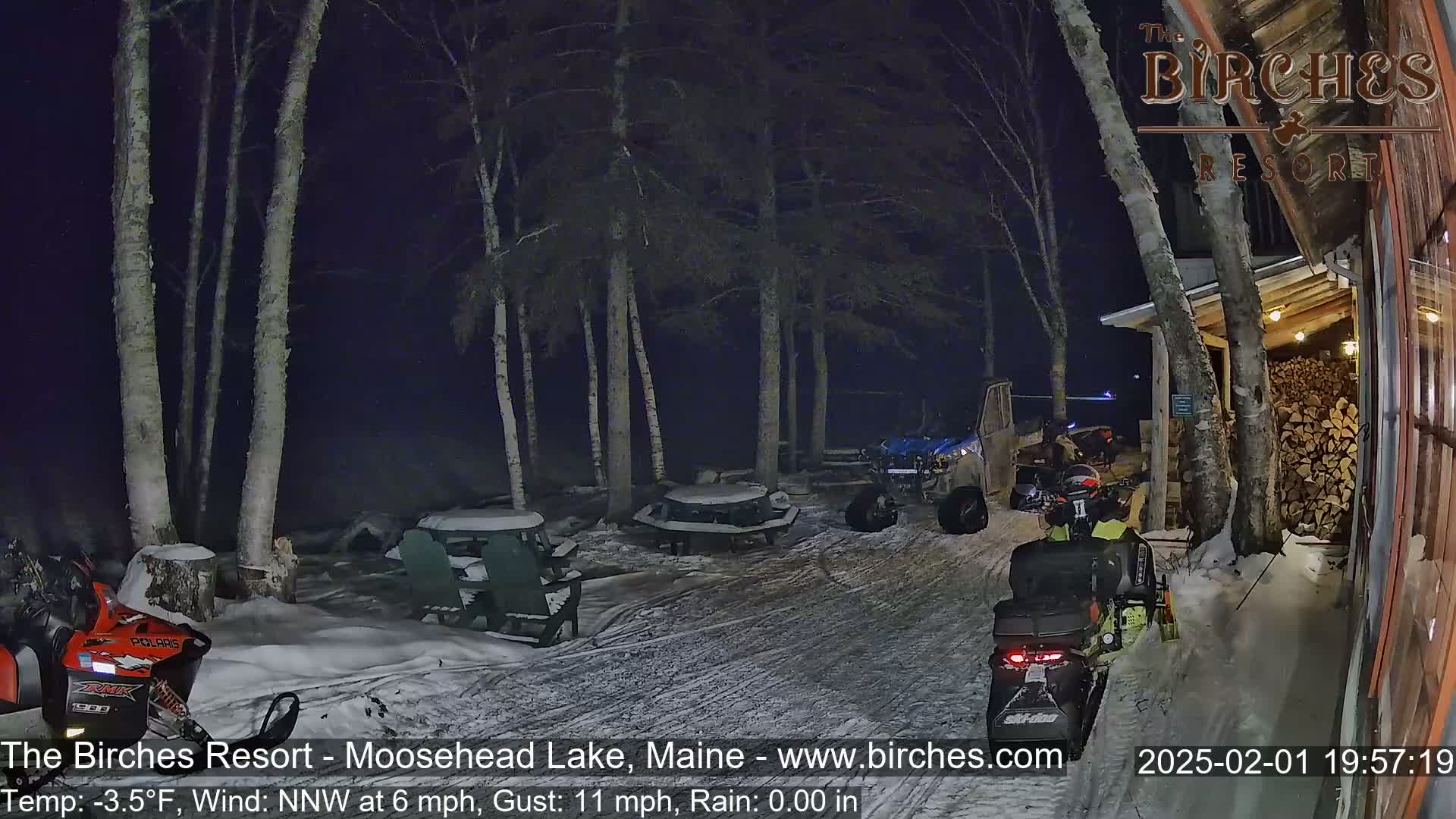 A nighttime scene shows snow-covered picnic tables, snowmobiles, and a woodpile near a building under a dark sky with light rain or snow.