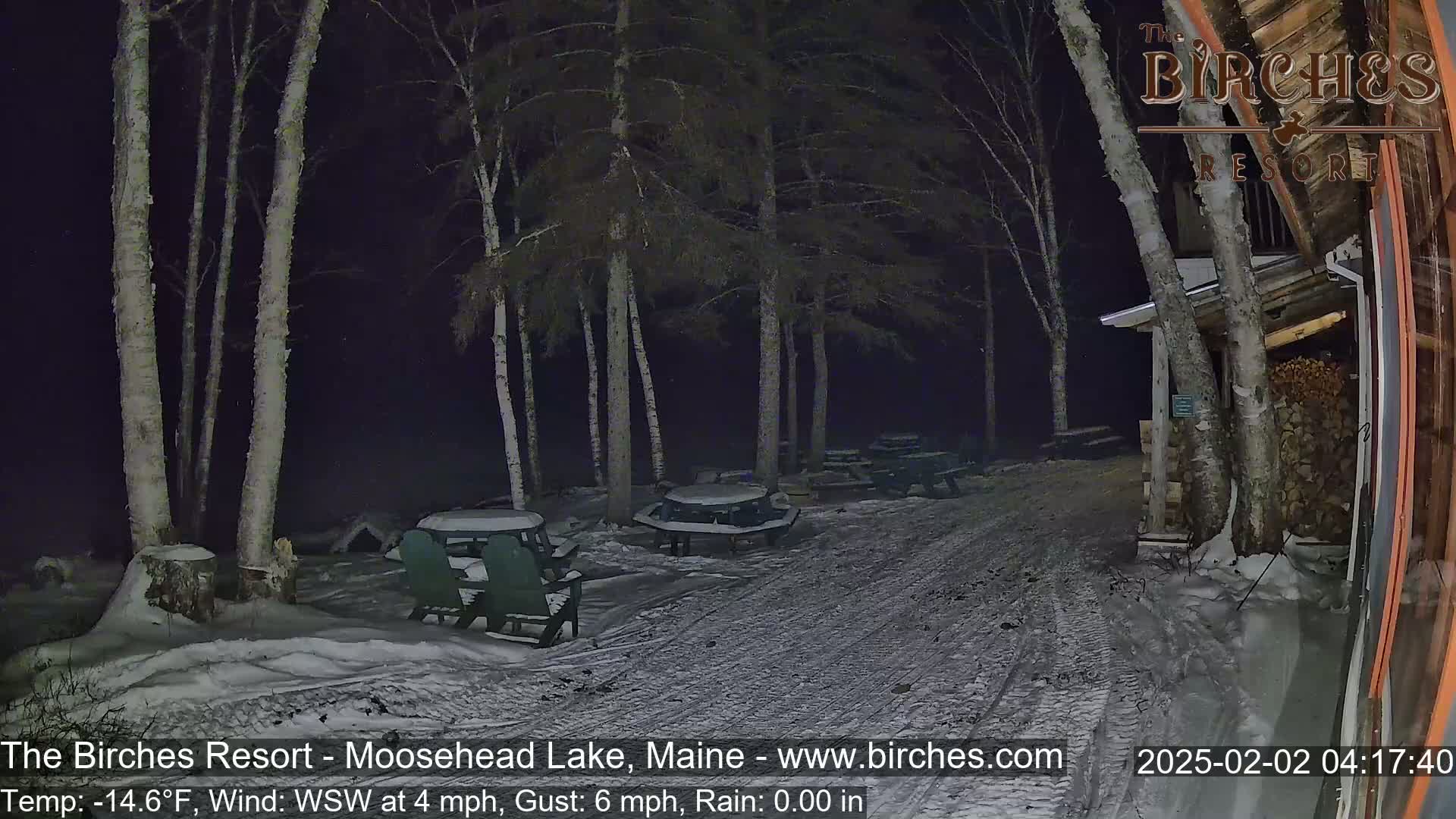 A snow-covered pathway leads to several picnic tables and chairs under a dark night sky, with a building visible in the background, and a slight wind and rain are present.