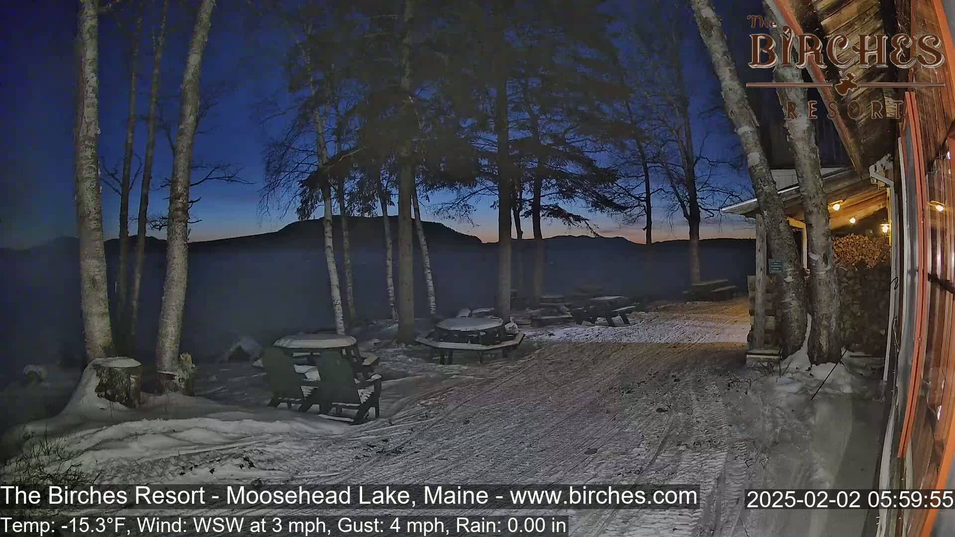 A snow-covered outdoor area with picnic tables and chairs is visible at twilight, under a dark blue sky with a calm lake in the background.