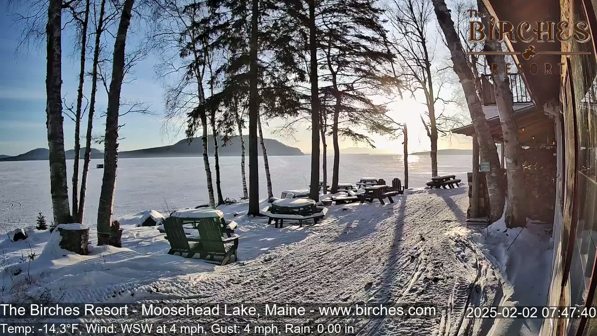 A snowy lakeside patio with picnic tables and chairs sits near a building, under a sunny, cold winter sky.