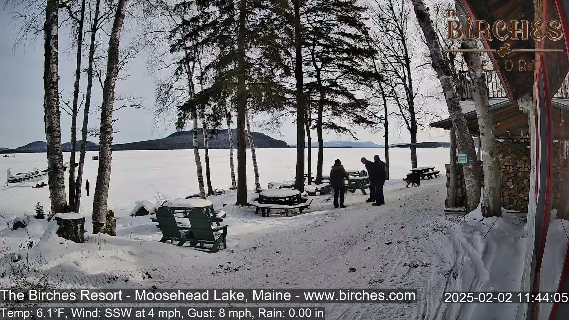 A snow-covered lakeside scene features several people near picnic tables and a building, with a frozen lake and distant hills under a clear sky.
