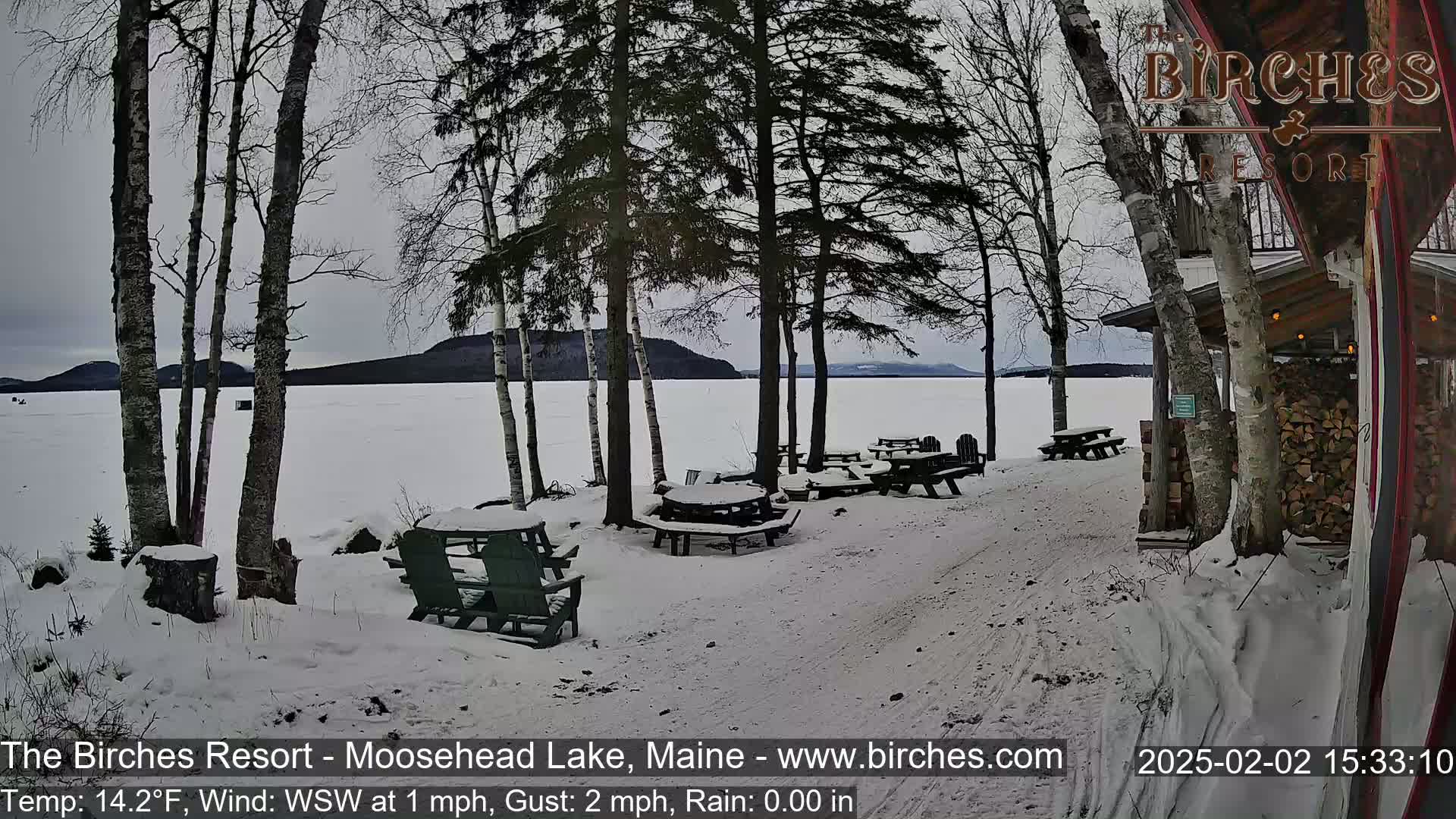 A snow-covered lakeside patio with picnic tables and chairs, under a cloudy sky, overlooks a frozen lake and distant mountains.